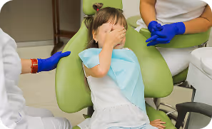 Young girl sitting in a dental chair covering her eyes while a dentist and assistant with blue gloves prepare to treat her.