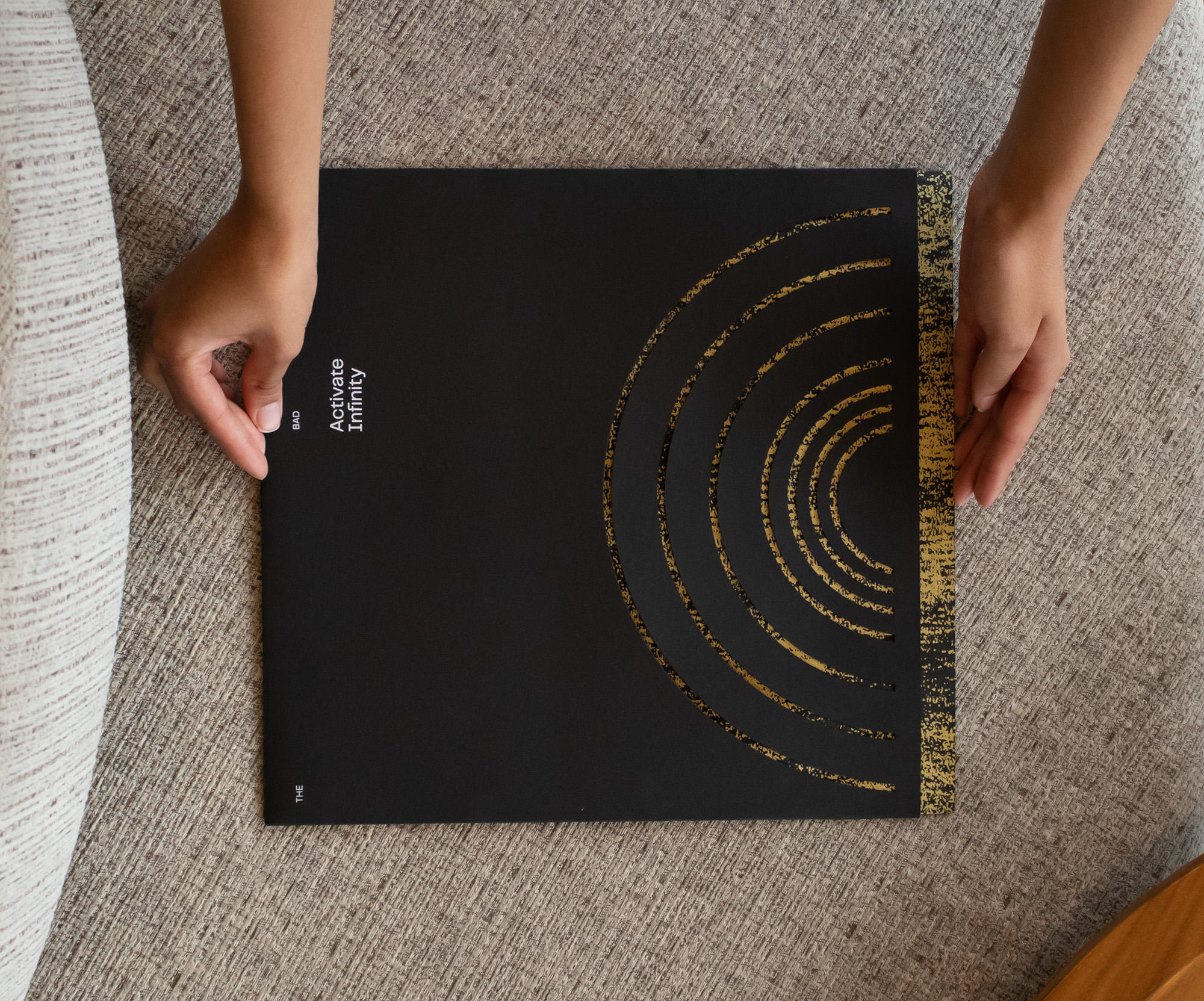 Two hands slide a gold patterned record sleeve into a black album jacket. Seven concentric semi-circles cut into the right side of the jacket allow the sleeve to be seen through the front. Text on the left side reads “The Bad Plus” and “Activate Infinity”.