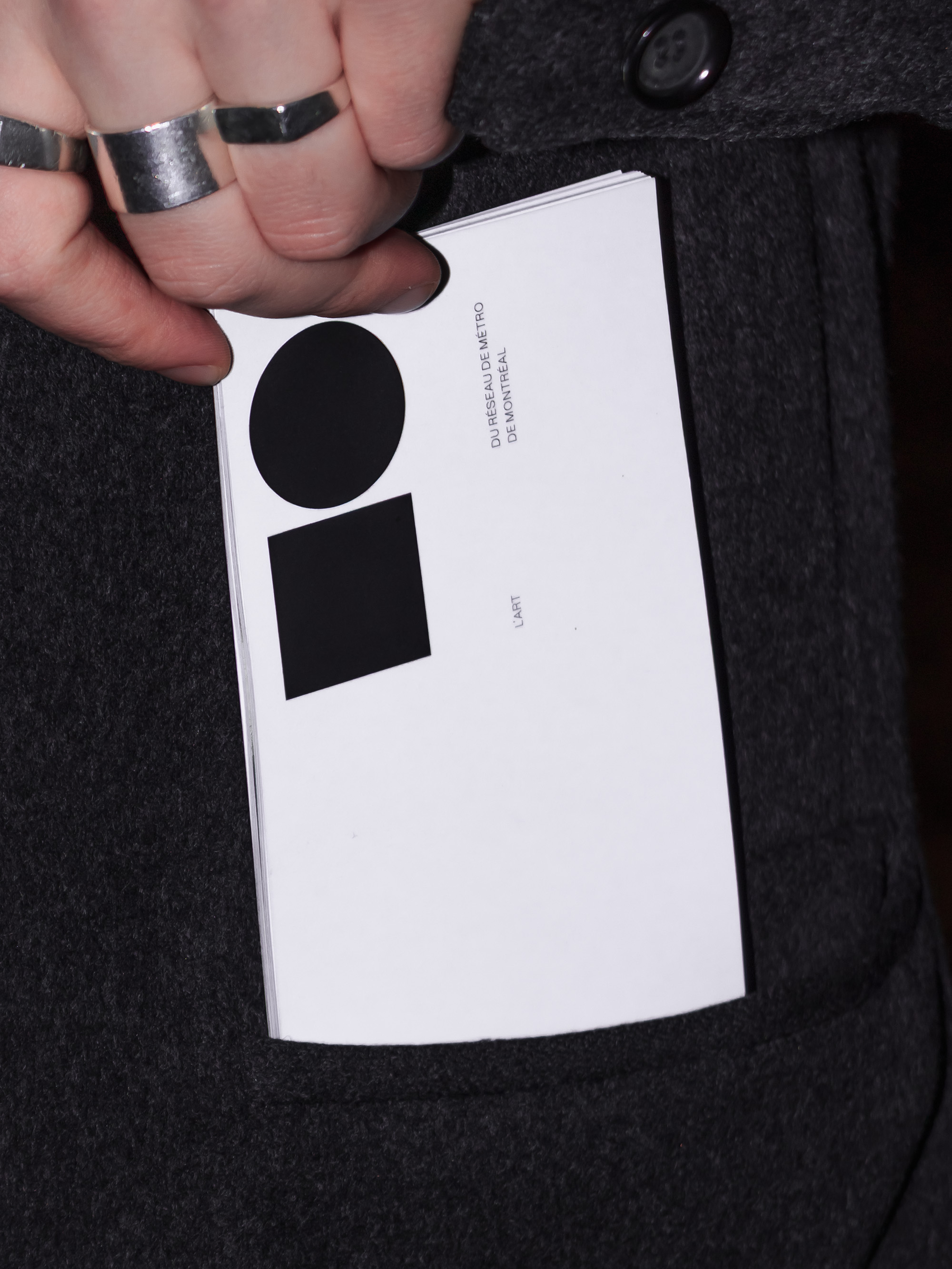 Closeup of a booklet being slid into a black coat pocket by someone wearing three silver rings. The cover prominently features a black square, underneath which is the word “L’Art”, next to a black circle, underneath which is the rest of the title: “du Réseau de métro de Montréal”.