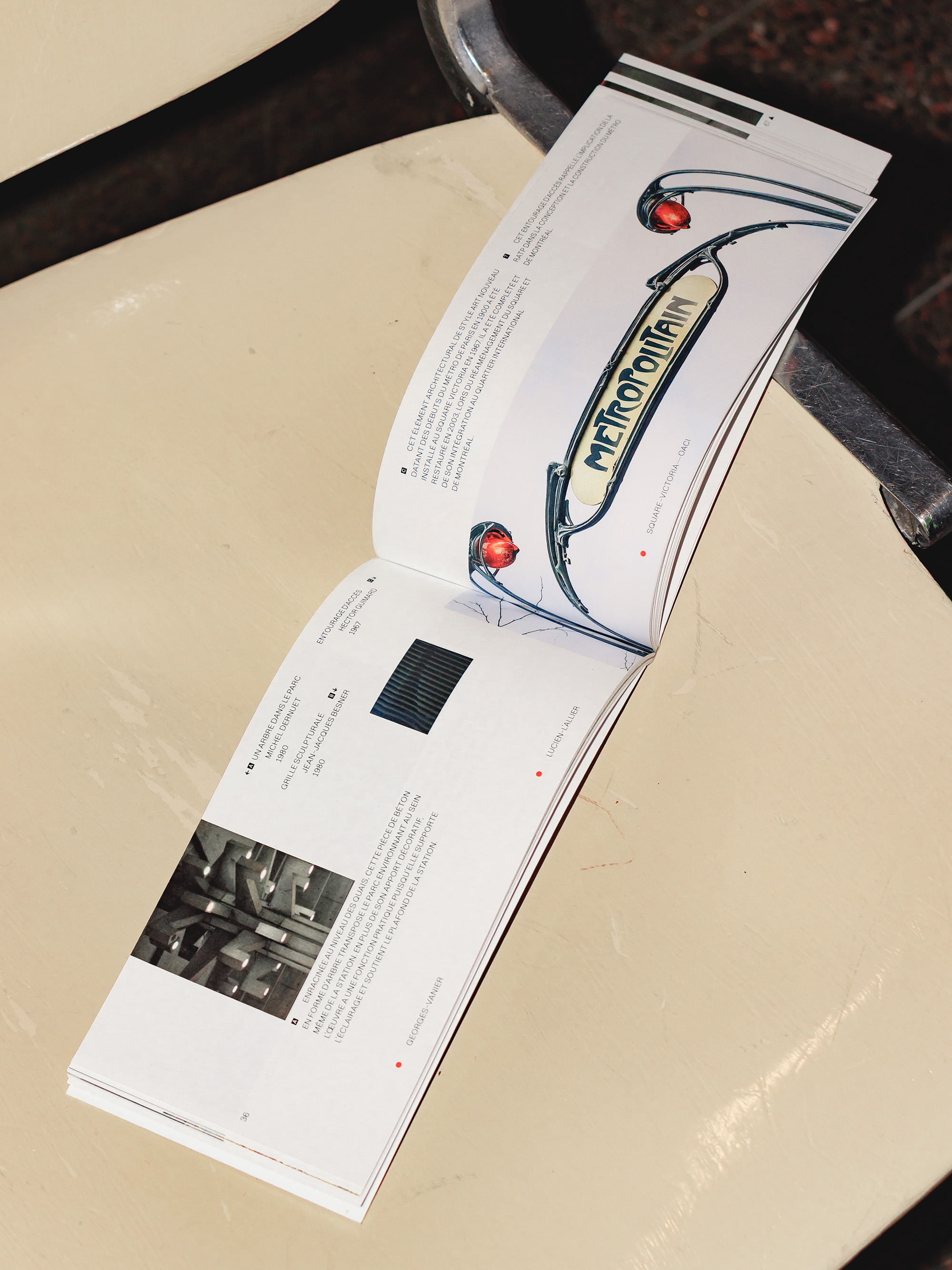 A booklet open on a beige train station seat, placed diagonally from the bottom-left to the top-right of the frame. Station names at the bottom edge of the spread are aligned with photos and descriptions of the public art pieces they house. 