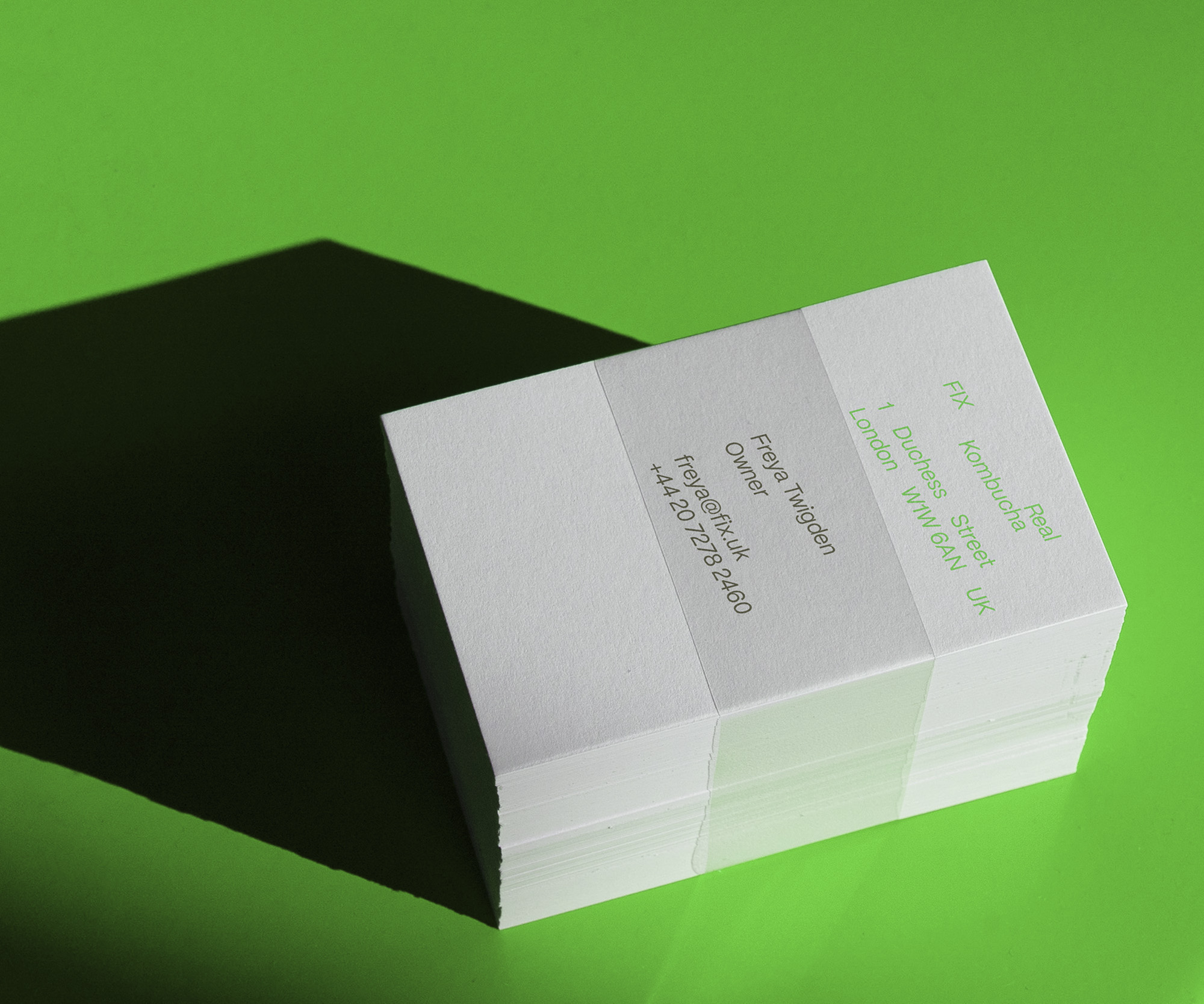 A stack of business cards on a bright green surface, casting a dark shadow. Diagonal, slanted text lists the company details and an employee’s contact information.