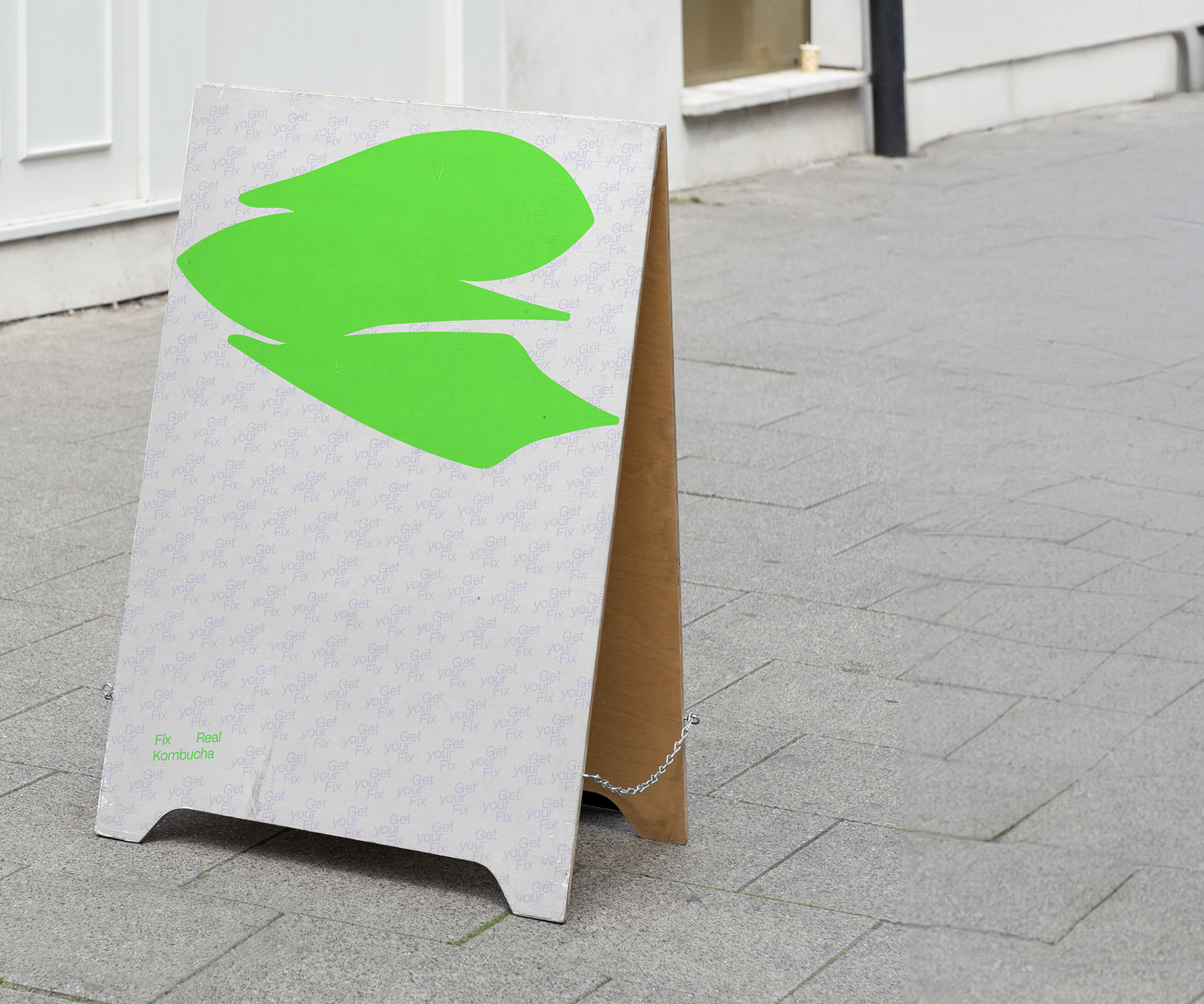A white a-frame sign on a sidewalk with a bright green shape at the top, pointing towards a nearby building. Repeating, soft purple text behind the shape reads “Get your Fix”, with “Fix Real Kombucha” in the bottom-left corner.