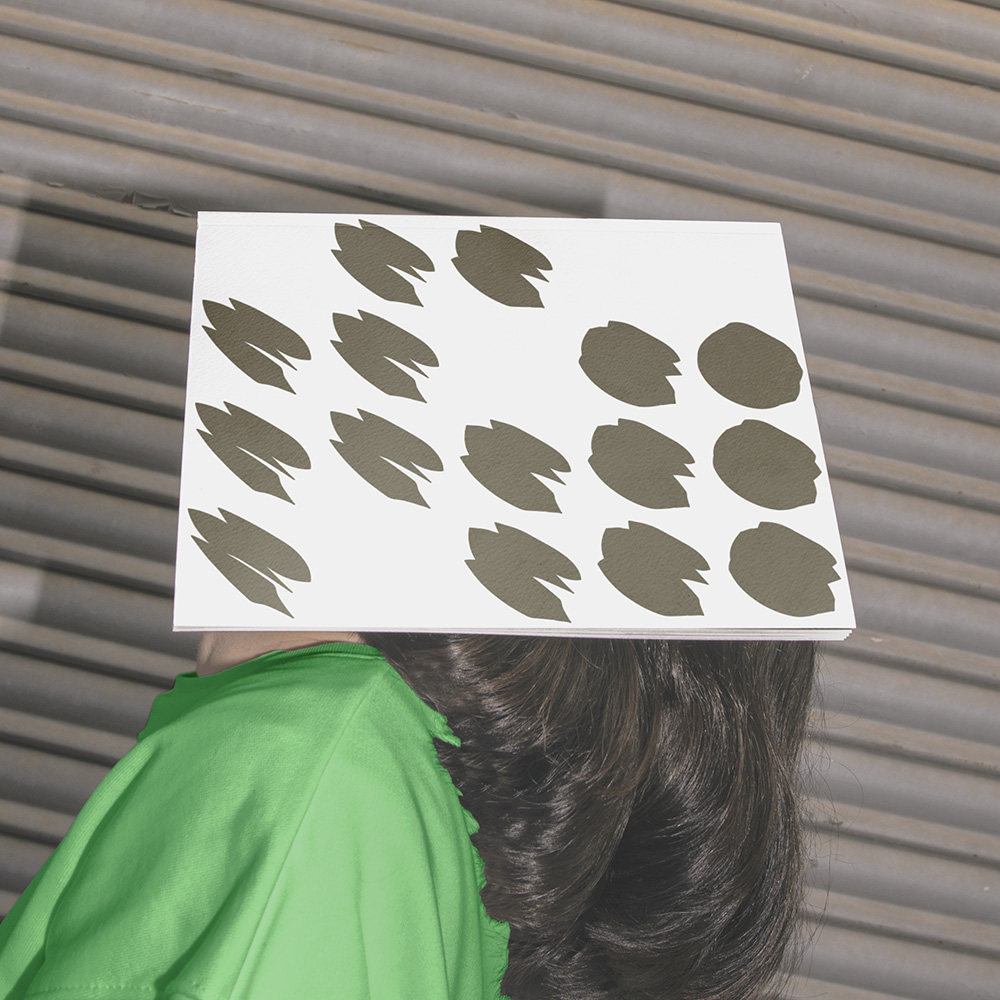 Side view of a person looking upwards with a white booklet covering their face. The cover of the booklet features a series of dark bronze shapes, progressing from soft and rounded to exclamatory in appearance. 