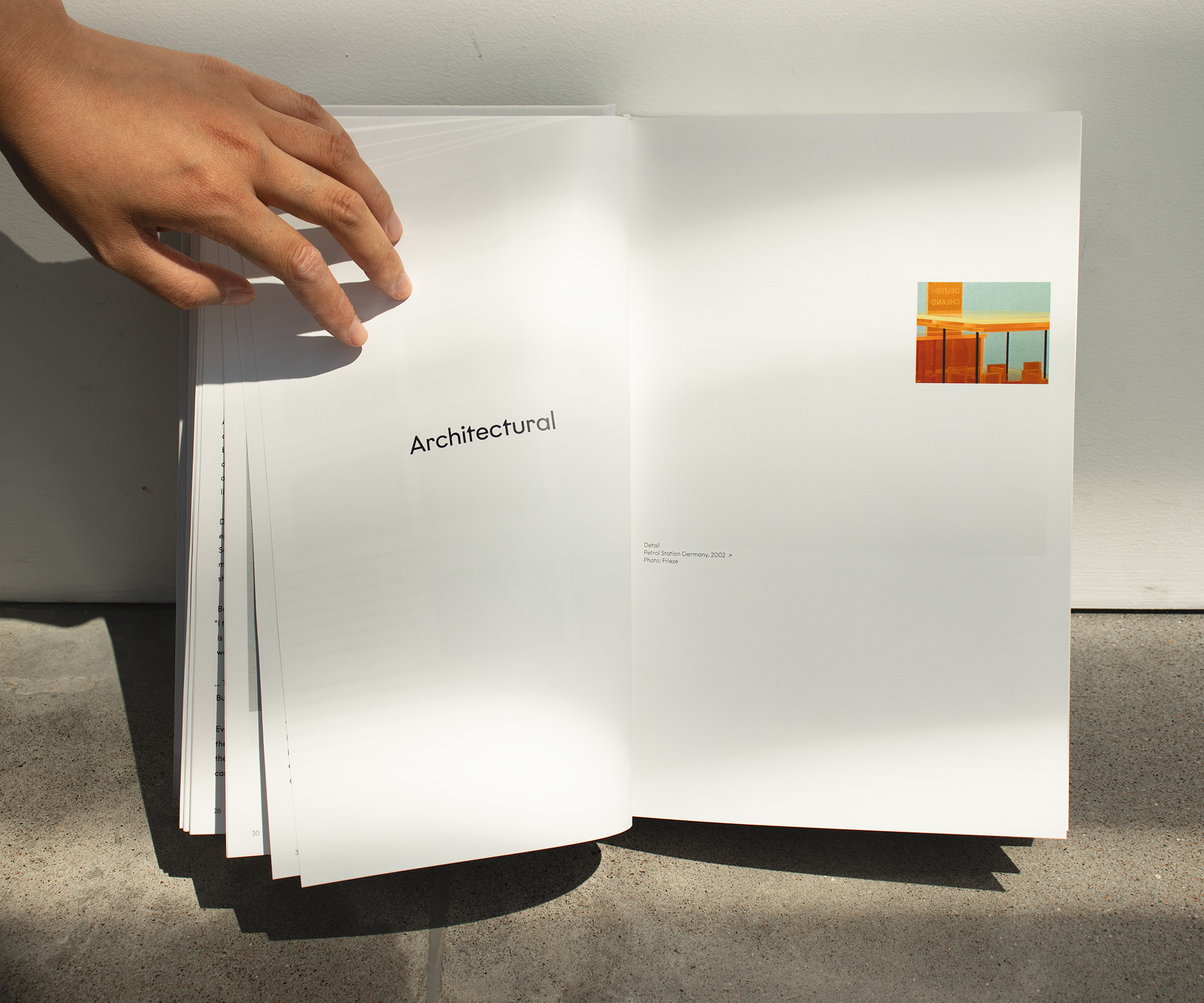 A book leaning against a white background on a polished concrete floor, held open by a hand from the top-left corner. Text on the left page reads “Architectural”, with a small, rectangular image on the right and a caption reading “Petrol Station Germany, 2002.”