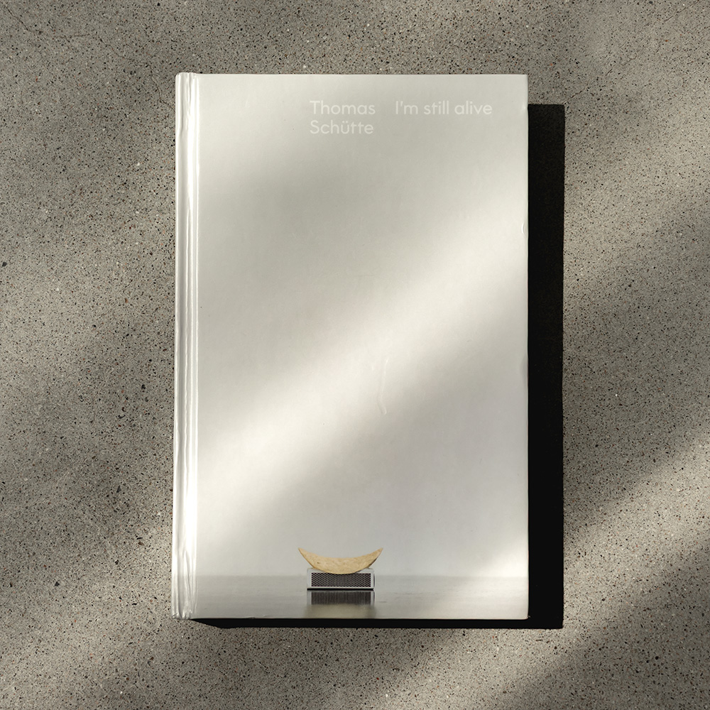 The front of a closed, portrait-shaped book on a concrete floor. The cover is a light grey, with Thomas Schütte’s “Pringle” (a single Pringle chip on a matchbox) at the bottom, aligned with the title, “Thomas Schütte:” I’m still alive” in two blocks of text at the top.