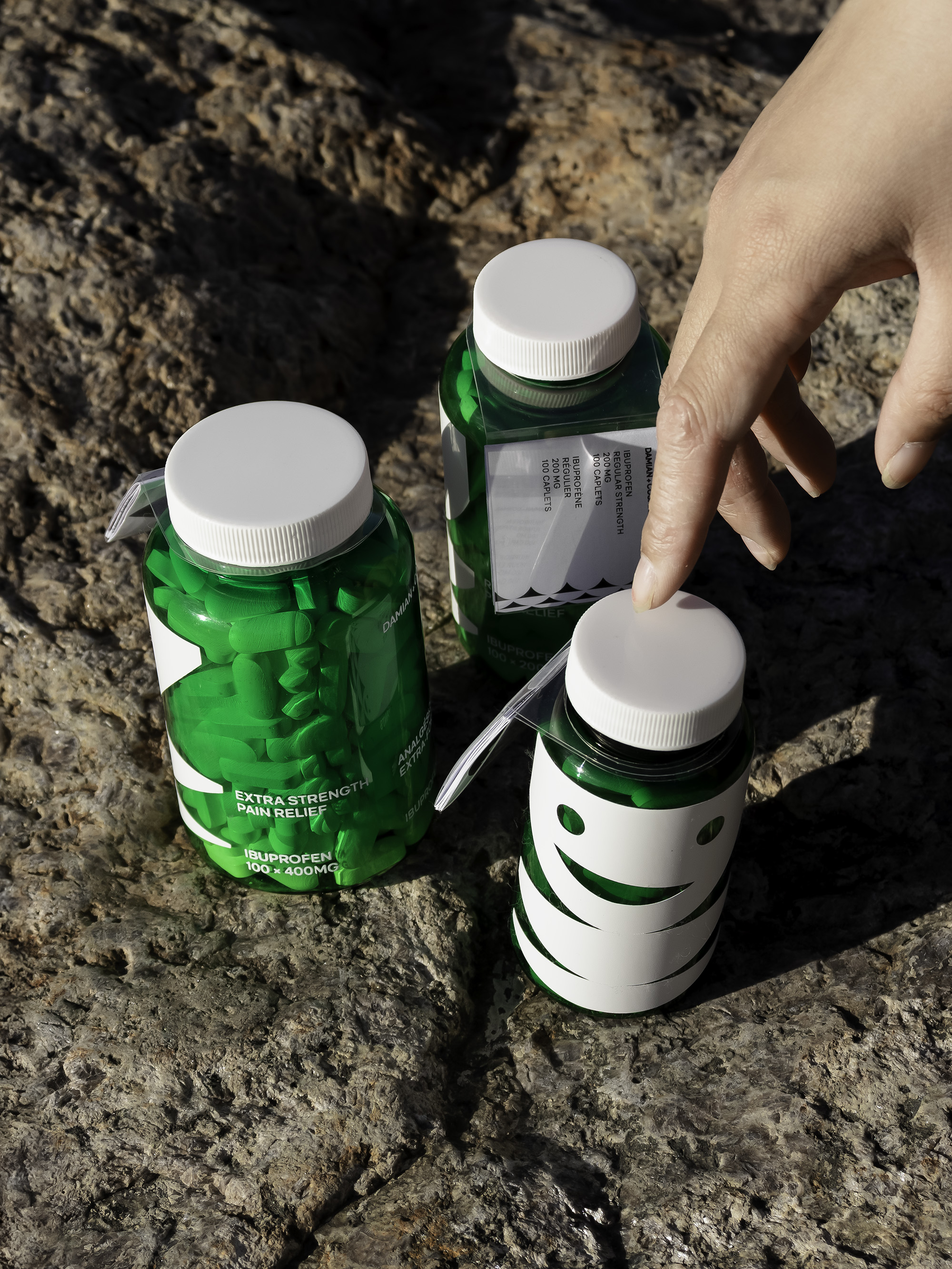 Three translucent green pill bottles with white text and geometric graphics sitting on a rocky surface. Tags are affixed around the necks of each bottle. A hand reaches for the largest from the top-left corner.