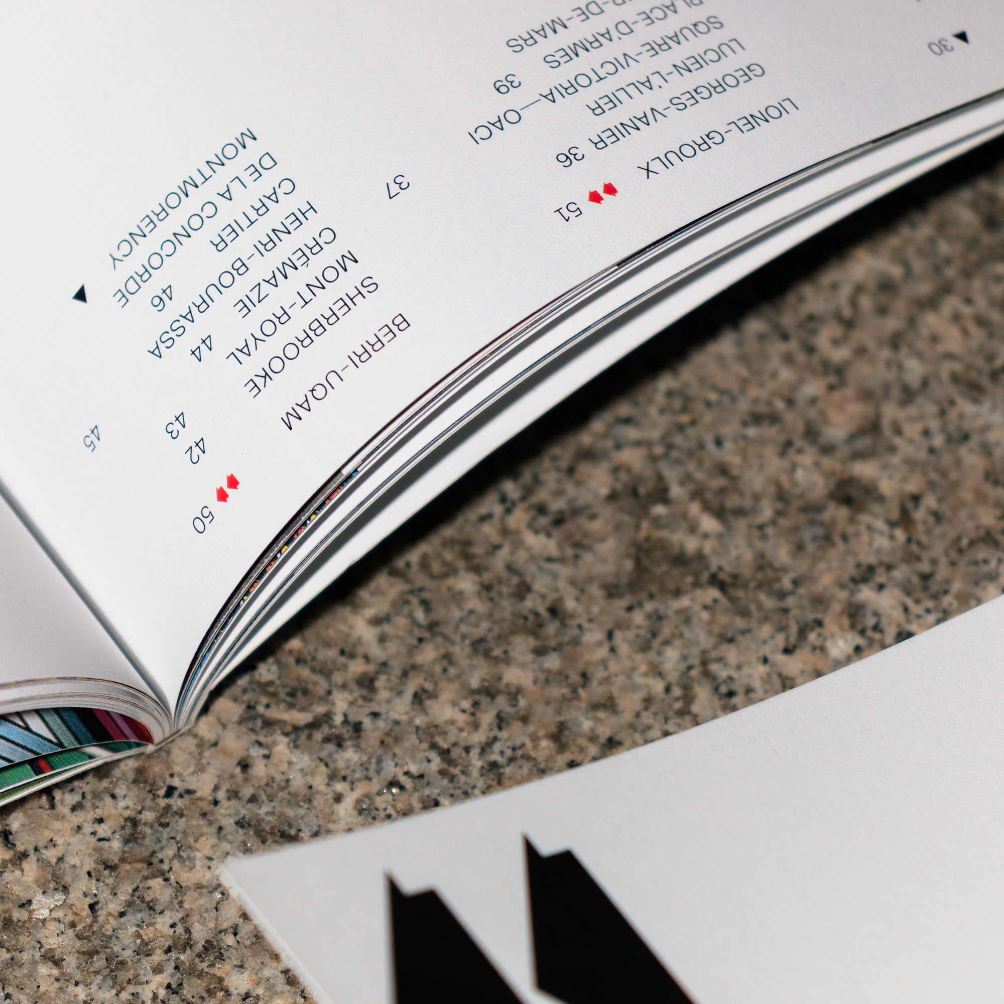 Closeup of the center of an open booklet with a chapters’ table of contents; a list of the orange line’s stations, placed on a granite background. Red arrows next to a station name indicate that its pages are found in another section, and a black triangle indicates the last station in the chapter. At the bottom-right, the corner of a second booklet is visible. 