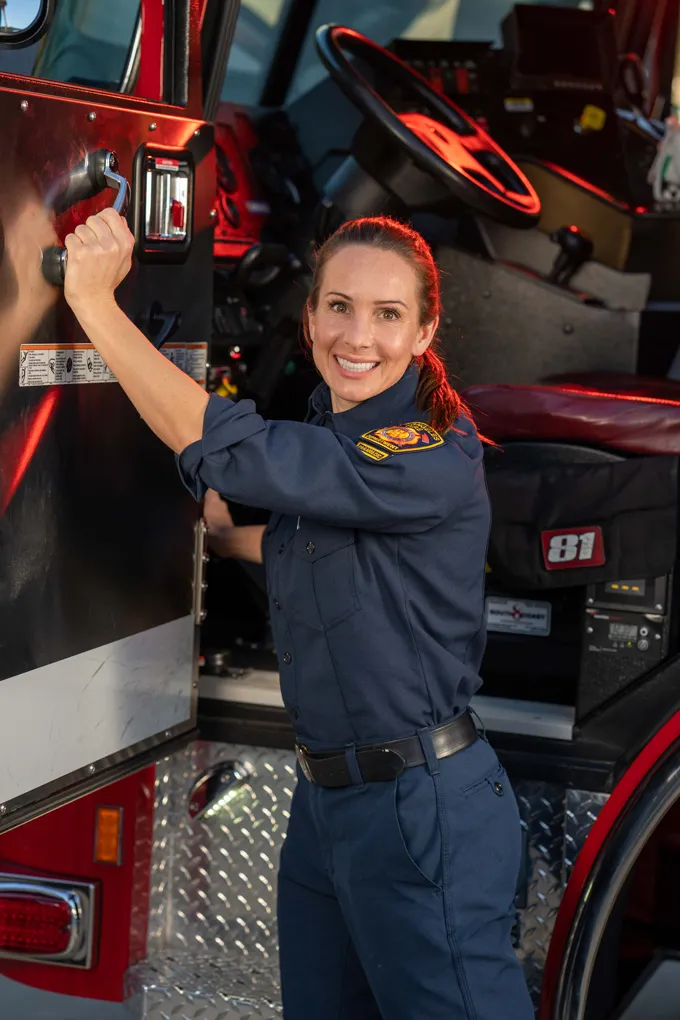 Smiling female firefighter in navy uniform opening the door of a fire truck.