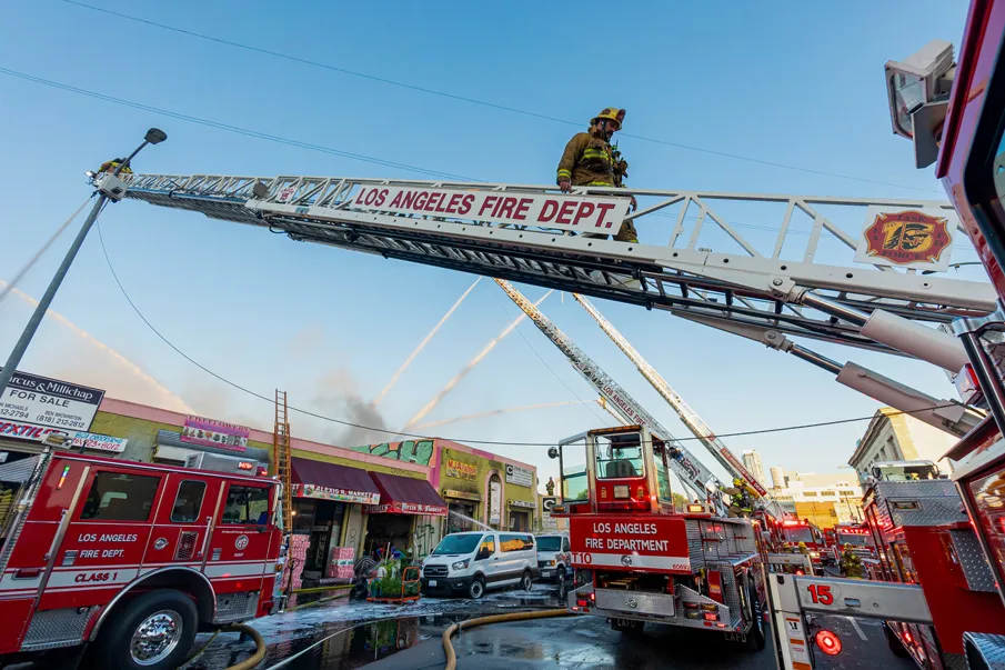 Los Angeles Fire Department trucks and firefighters responding to a fire at a commercial building with water hoses and ladders extended.