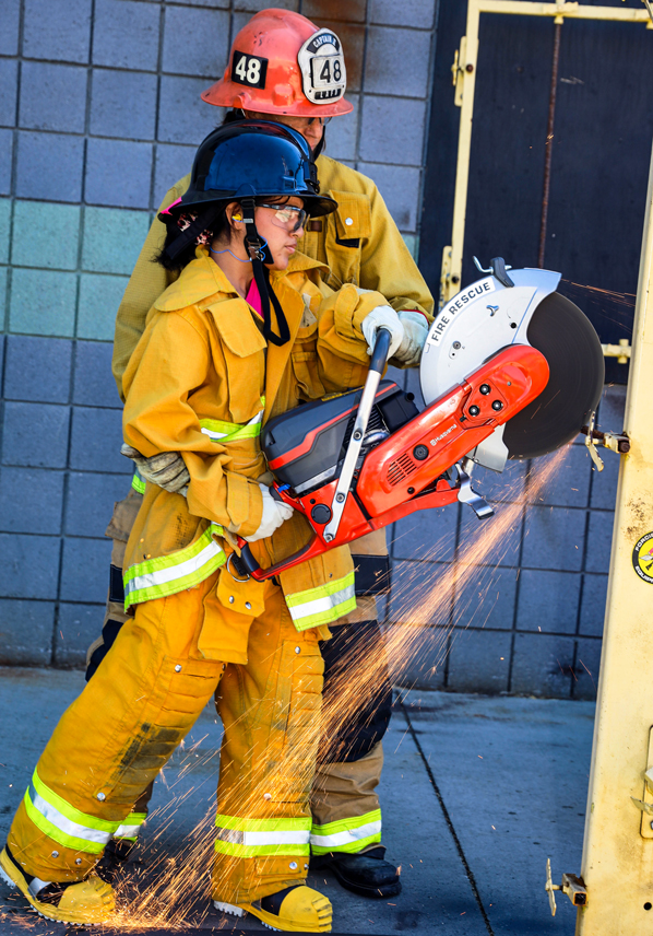 Camp Spark student wearing turnouts using a chainsaw to cut through a wooden wall.