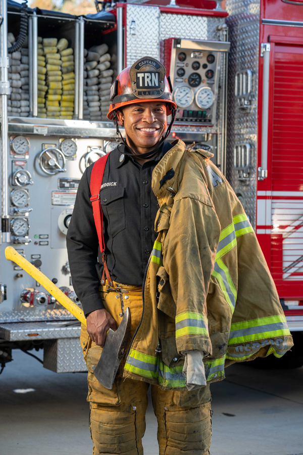 Male firefighter in helmet and uniform holding axe and yellow protective jacket, standing in front of a fire truck with equipment.