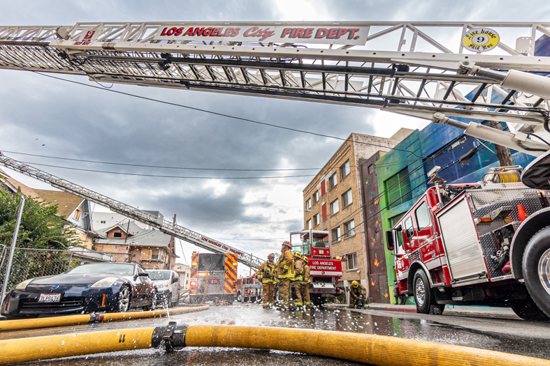 Los Angeles City firefighters in gear working near fire trucks and hoses on a wet street under a cloudy sky.