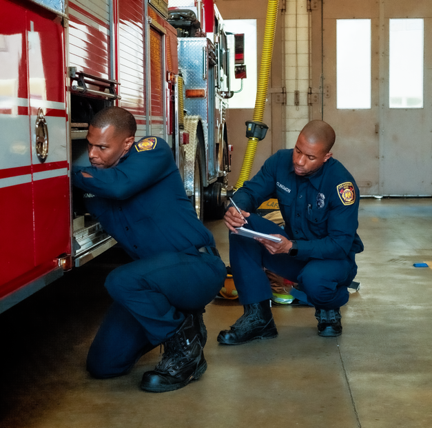 Two firefighters in uniform inspecting a red fire truck inside a fire station, one checking a compartment and the other taking notes.