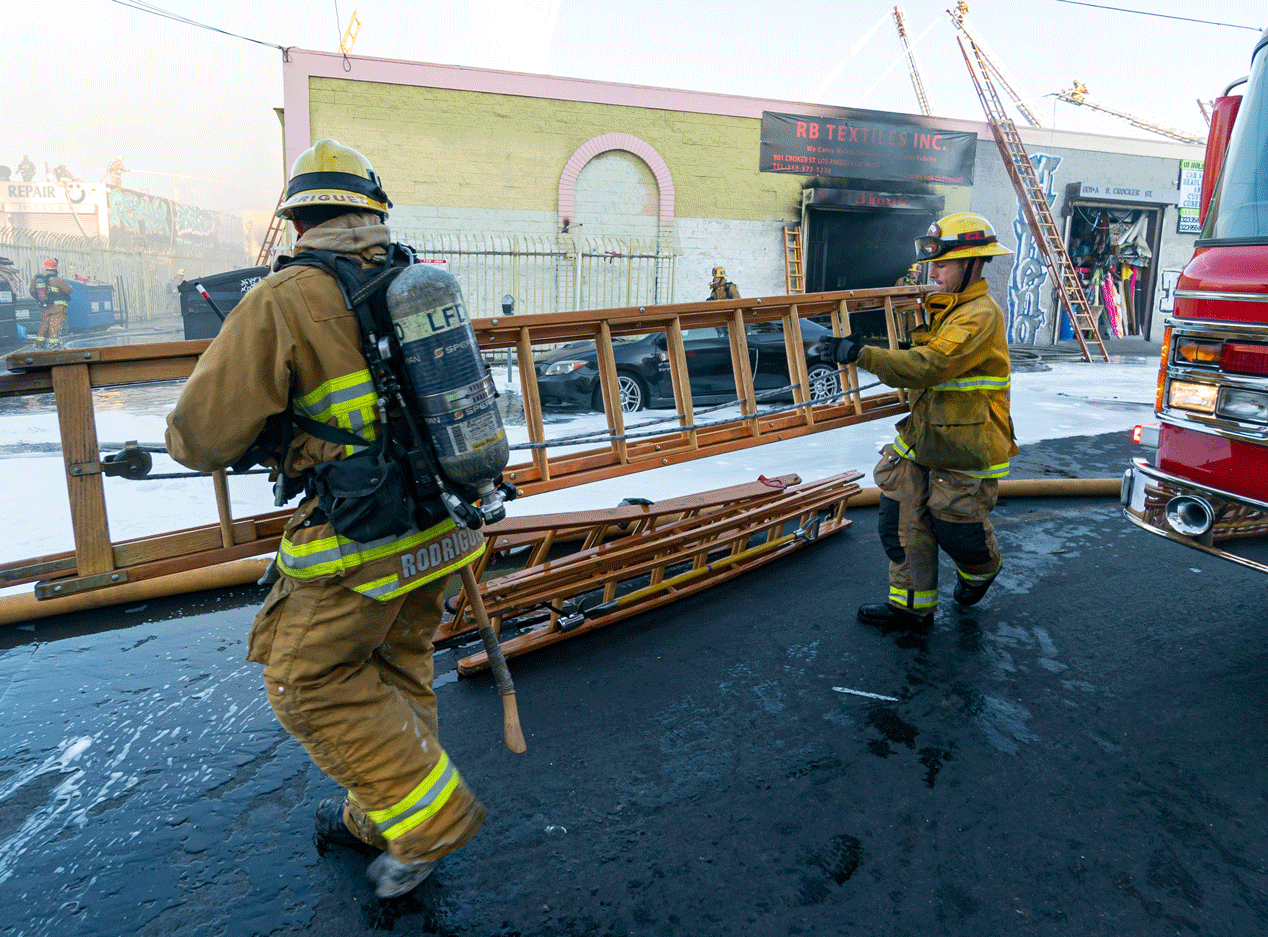 Two firefighters in full gear carrying a wooden ladder near a firetruck.