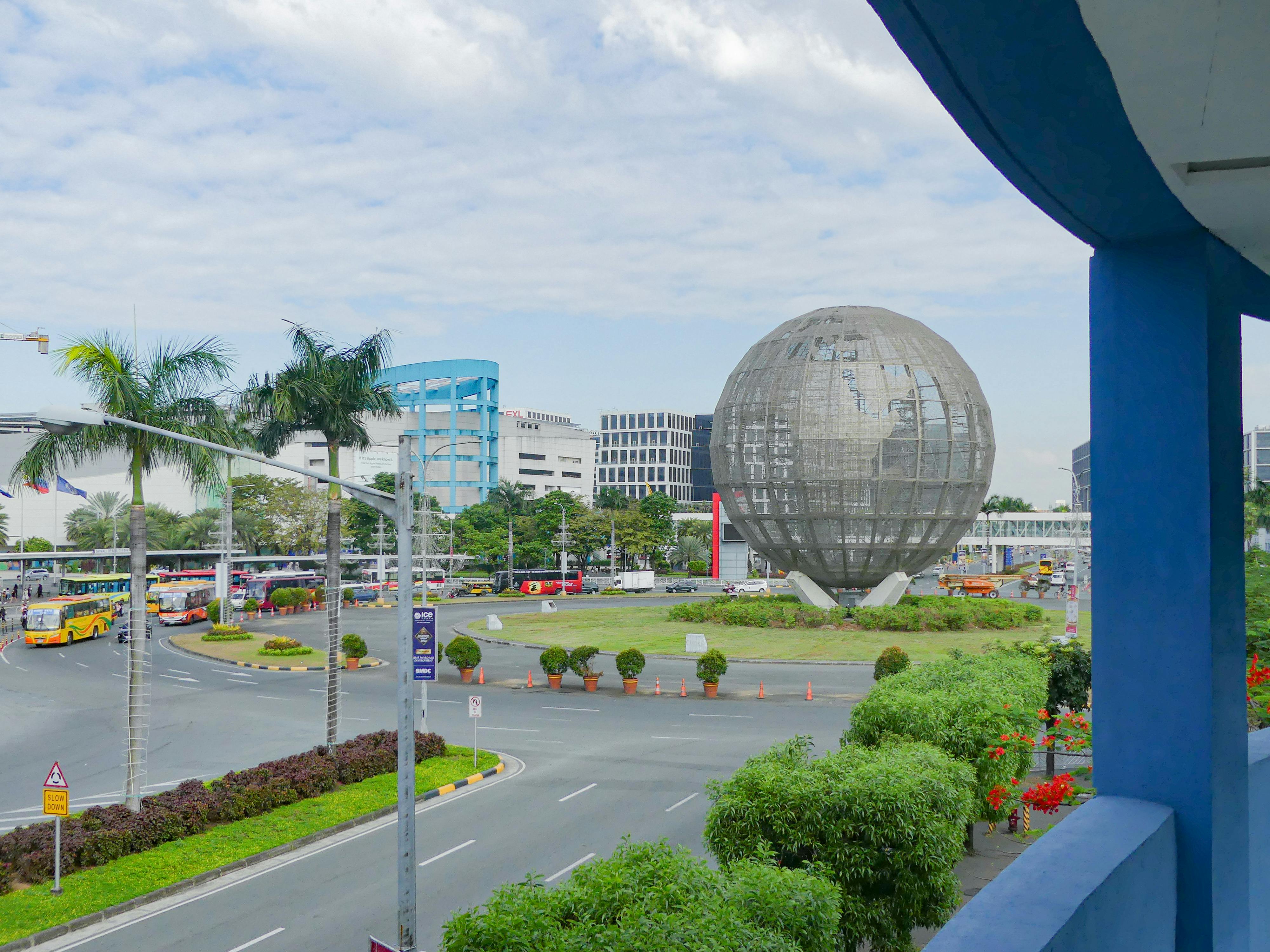 Urban roundabout with a large metal globe sculpture surrounded by greenery and buses in the background.