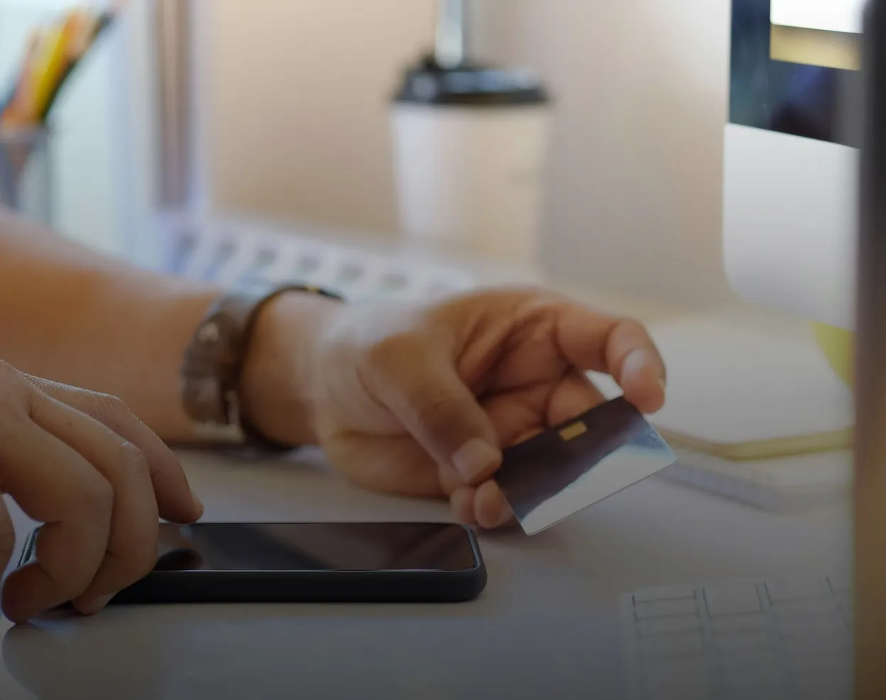 Close-up of a person holding a credit card in one hand and using a smartphone on a desk, with a computer monitor and notebook blurred in the background.