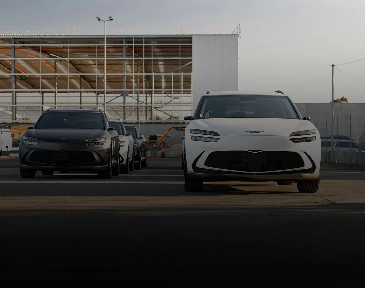 Line of modern cars parked outdoors facing forward with a white car in front and several dark-colored cars behind it, next to a large industrial building with metal framework.