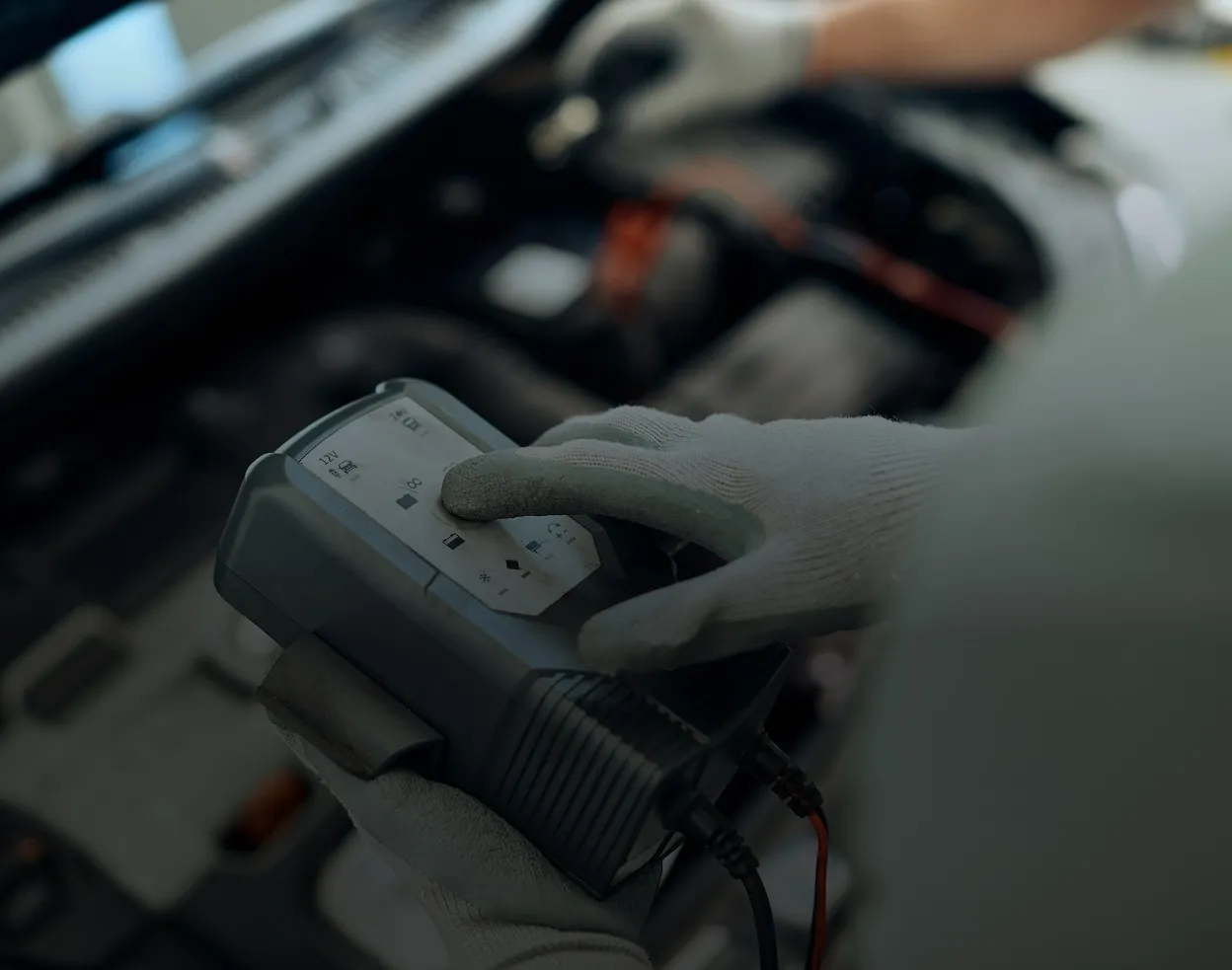 Close-up of a person wearing gray gloves using a digital automotive diagnostic tool, with a blurred car engine in the background.