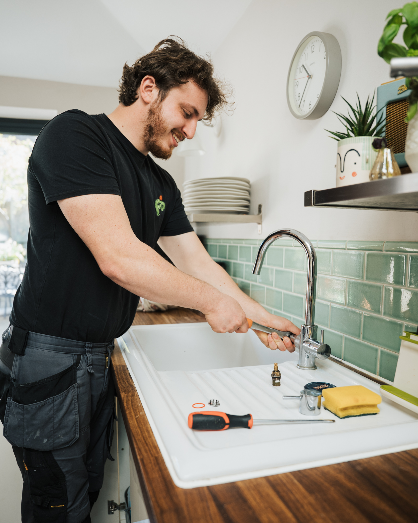 Plumber in a black T-shirt tightening a kitchen mixer tap with a wrench, tap parts on the white sink drainer