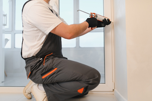 Worker in grey overalls kneeling to apply sealant with a gun along a window frame