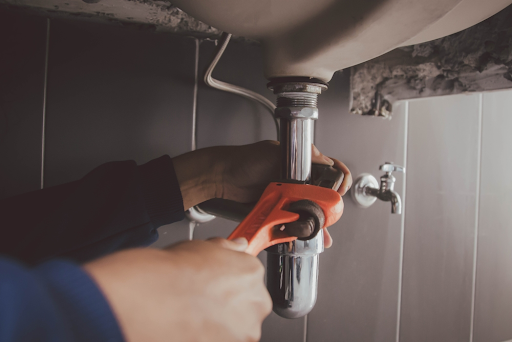 Hands using an orange adjustable wrench on a chrome bottle trap under a basin