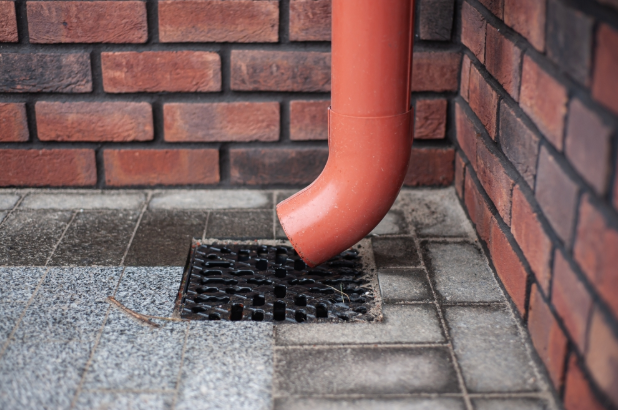 Orange downpipe running into a square drain grate on paving beside a brick wall