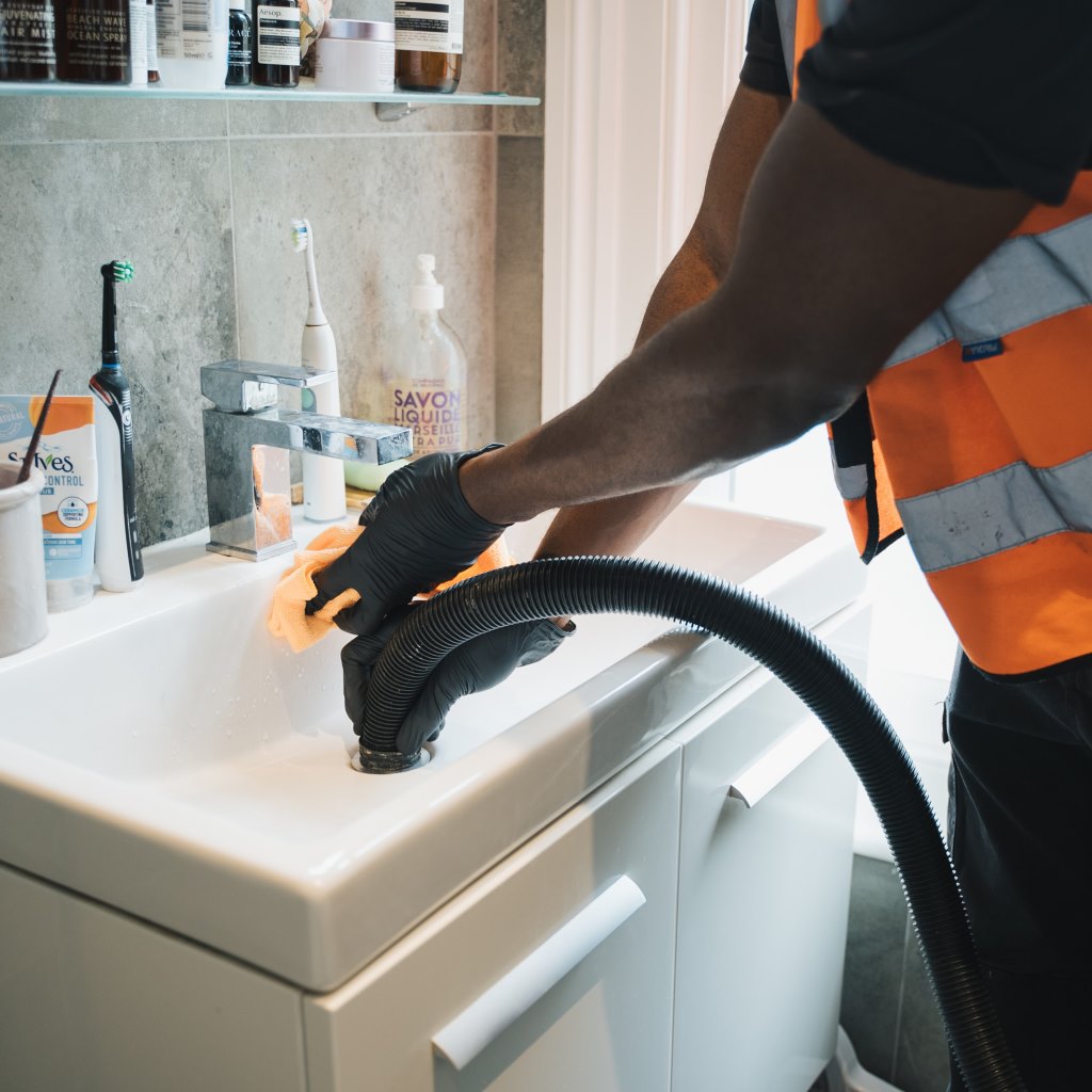 Worker in hi-vis vest using a black suction hose on a bathroom basin drain