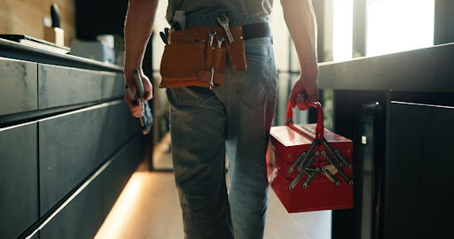 Tradesperson with a leather tool belt carrying a red cantilever toolbox through a modern kitchen