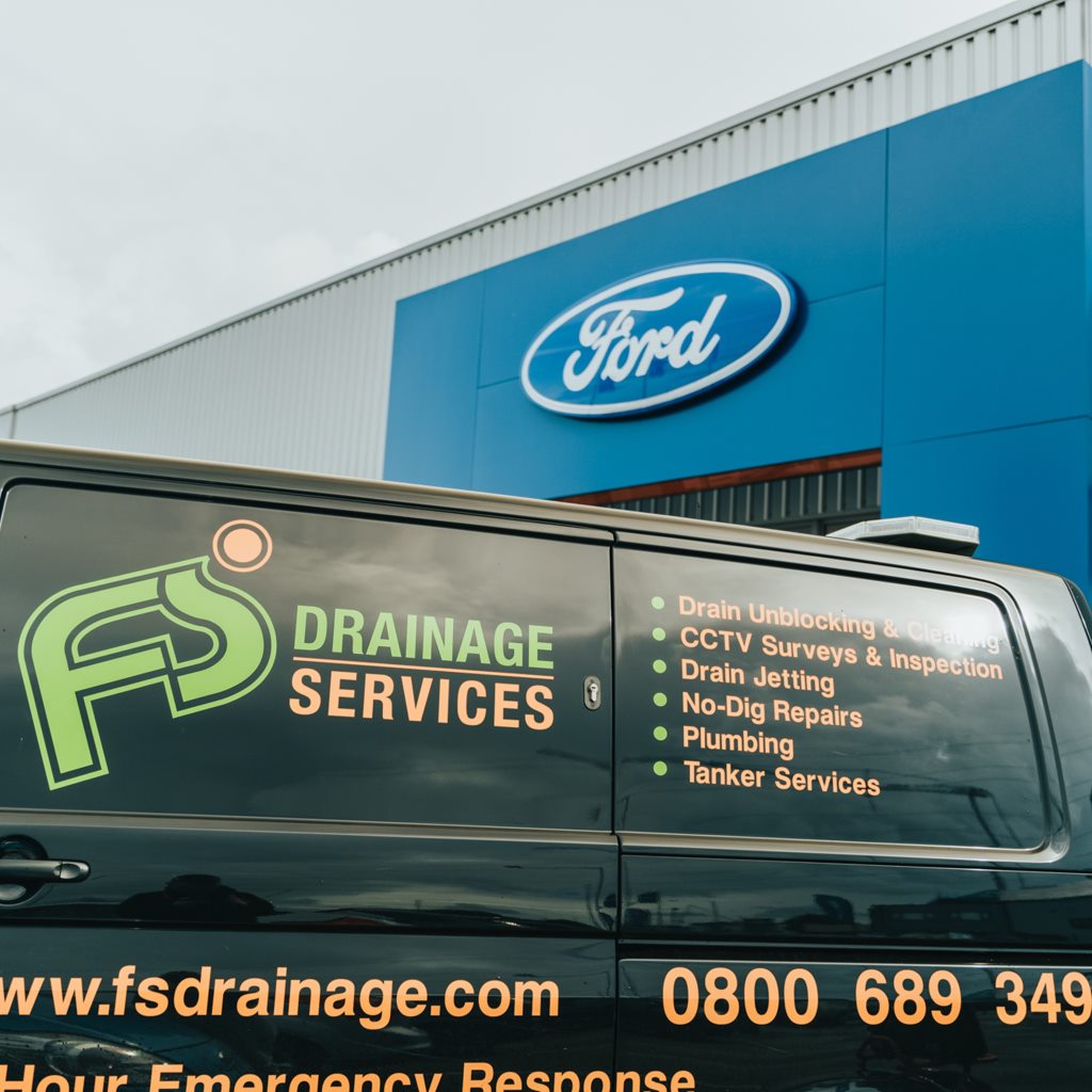 Black drainage van with a list of services on its side parked by a Ford building