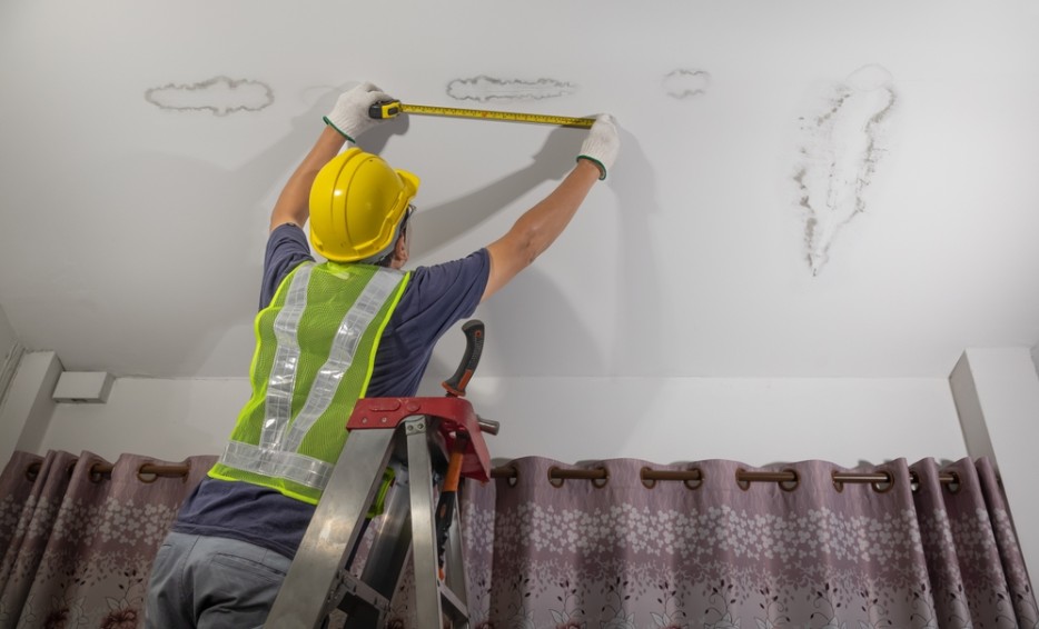 Worker in a hard hat on a ladder measuring water stains on a ceiling with a tape