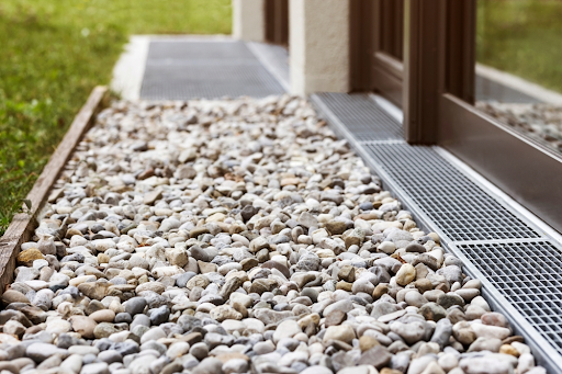 Pebble-filled drainage strip beside a building with metal grated channel drains along the glass doors