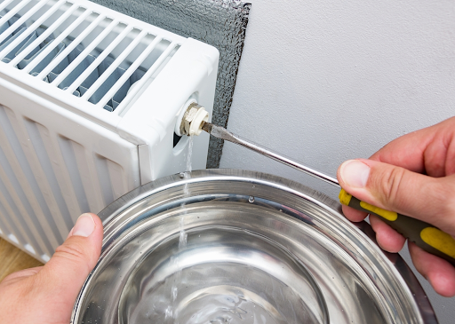 Hands bleeding a white radiator with a screwdriver while water drips into a steel bowl