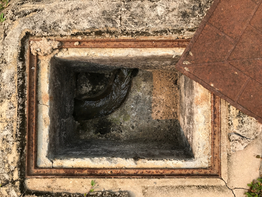 Open brick inspection chamber with a rusty frame and its cover lifted, showing the channel at the bottom