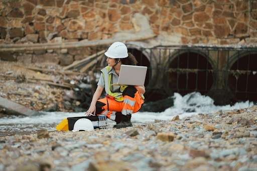 Engineer in a hard hat with a laptop crouching by a stream below large stormwater outfall pipes