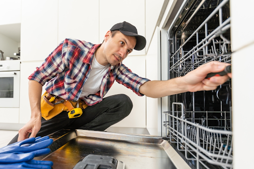Repairman in a cap and checked shirt using a screwdriver inside an open dishwasher
