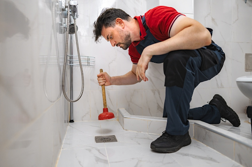 Plumber crouching with a red plunger beside a floor drain in a walk-in shower