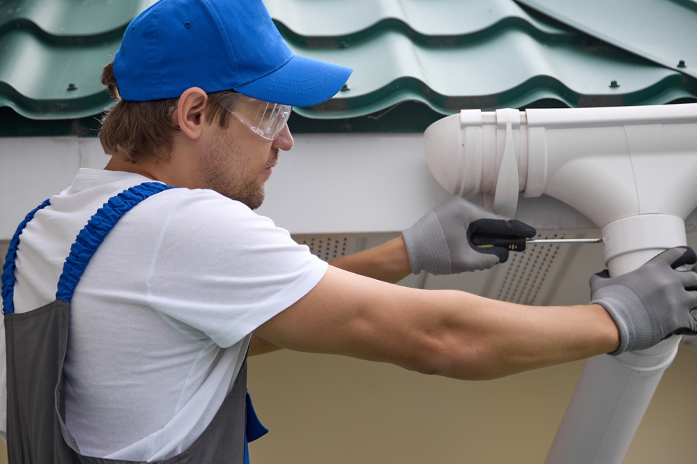 Worker in a blue cap and safety glasses fitting a white downpipe under a roof gutter