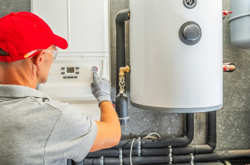 Engineer in a red cap pressing a button on a boiler control panel beside a hot water cylinder