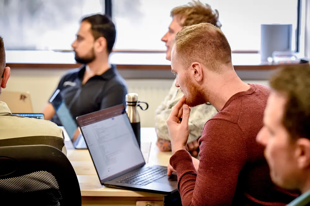 A group of men attentively listening and one man focused on a laptop screen in a meeting room.