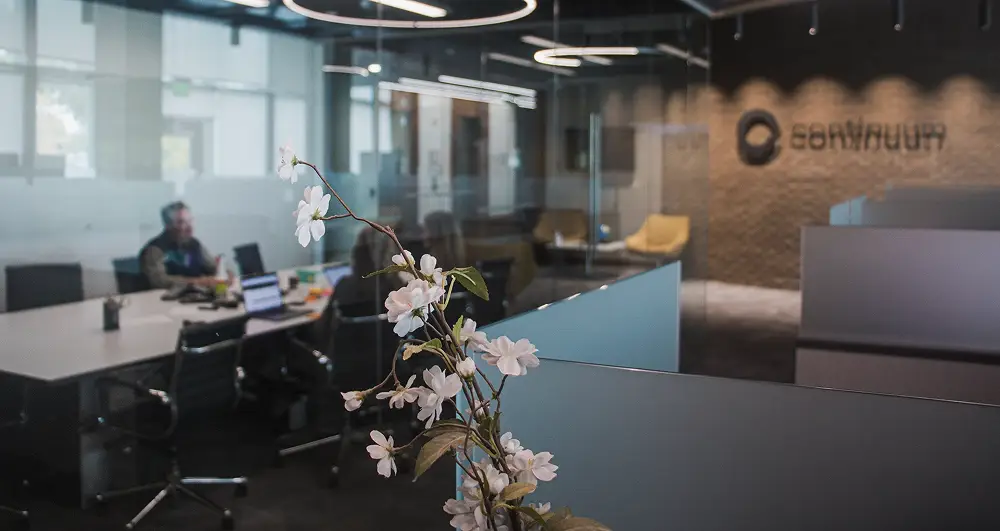 Modern office with glass walls, white flowers in foreground, and people working at a conference table.