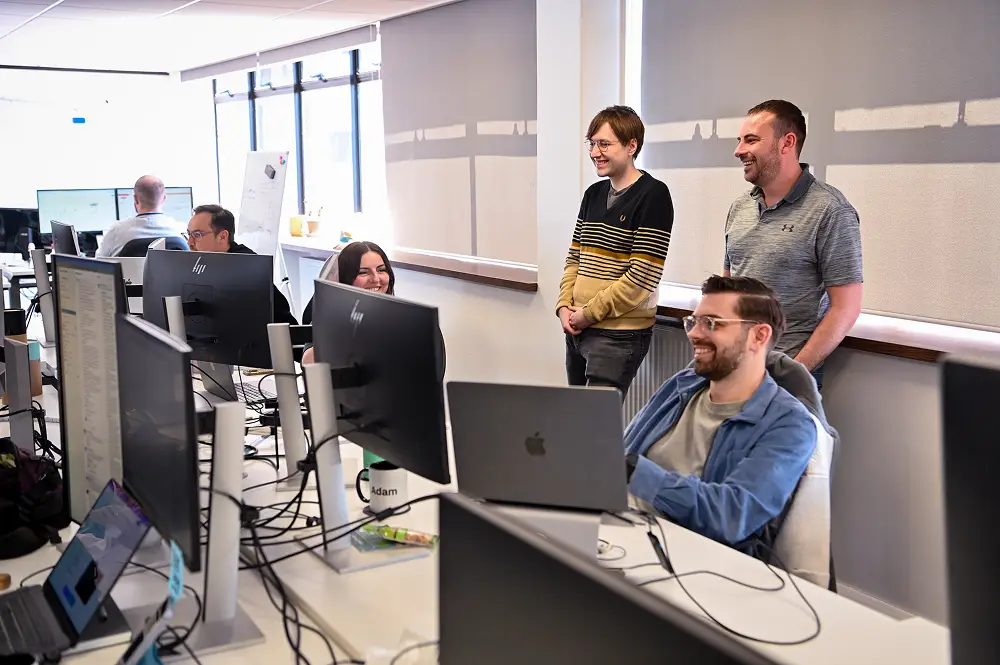 Group of five people in an office with multiple computer monitors, working and smiling together.