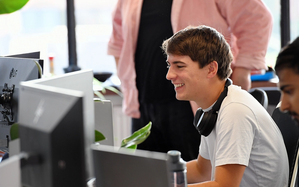 Smiling young man with headphones around his neck working at a computer in a bright office.