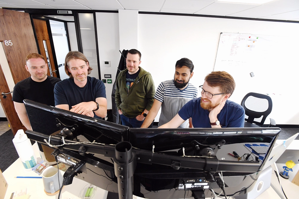 Five men gathered around dual computer monitors in an office, collaborating on a project.