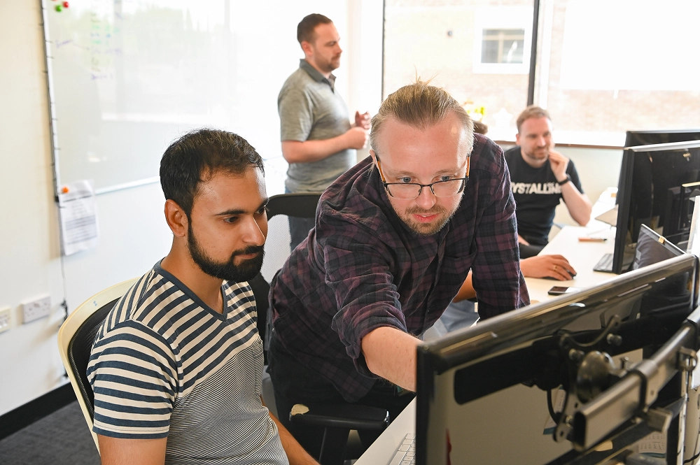 Two men focused on a computer screen while two others work and talk in the background office.