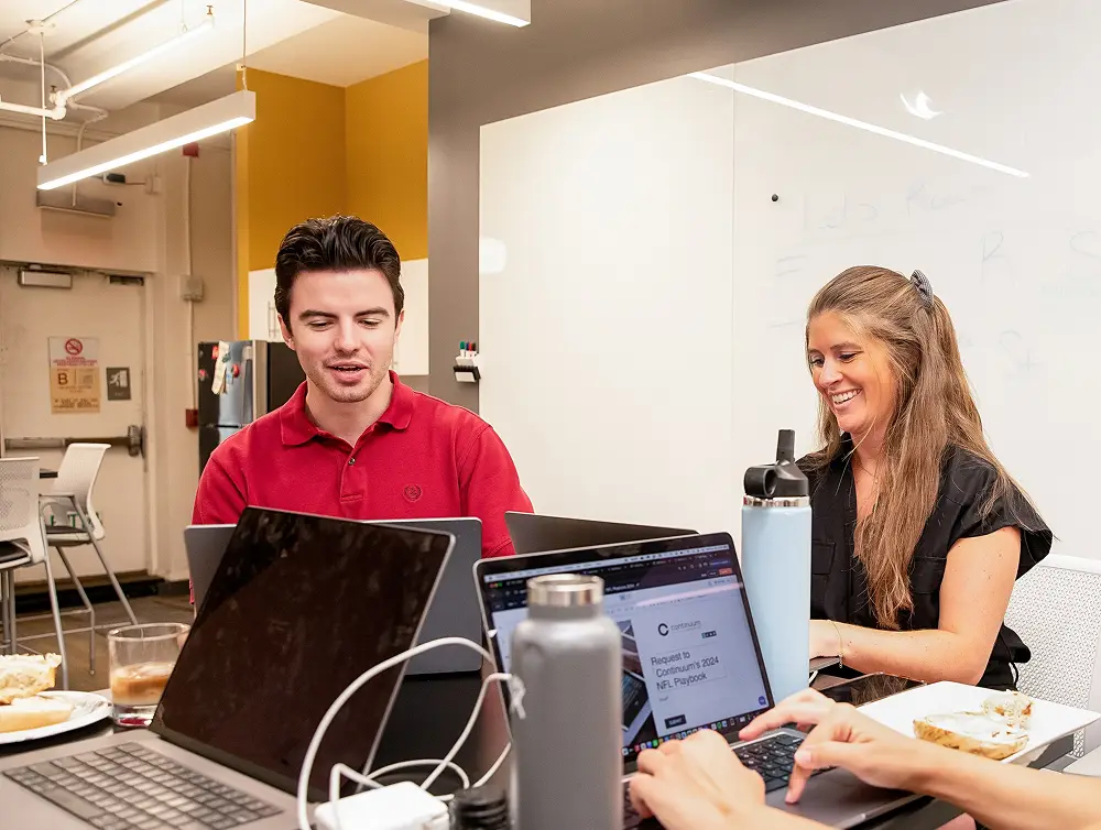 Two colleagues in an office meeting, one man in red shirt and a smiling woman, both using laptops.