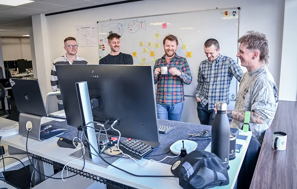 Five men laughing and chatting around desks with computers in a modern office space.