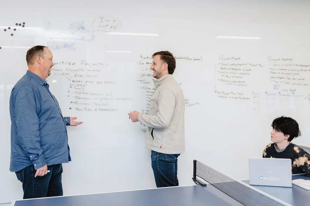 Two men discuss project plans on a whiteboard while a woman watches at a laptop near a ping-pong table.