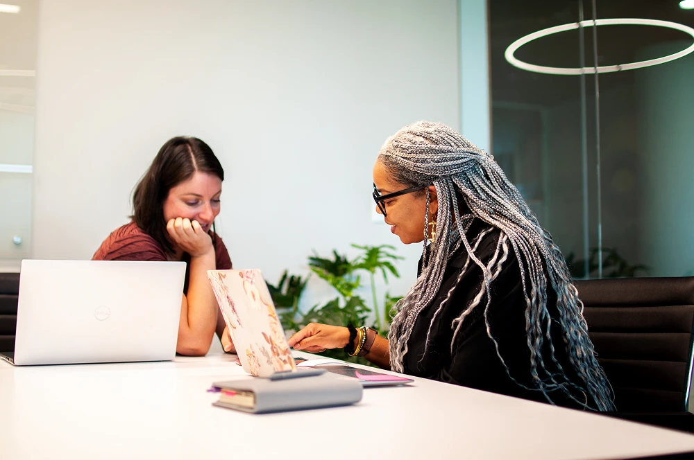 Two women working together at a table with laptops and documents in a modern office setting.