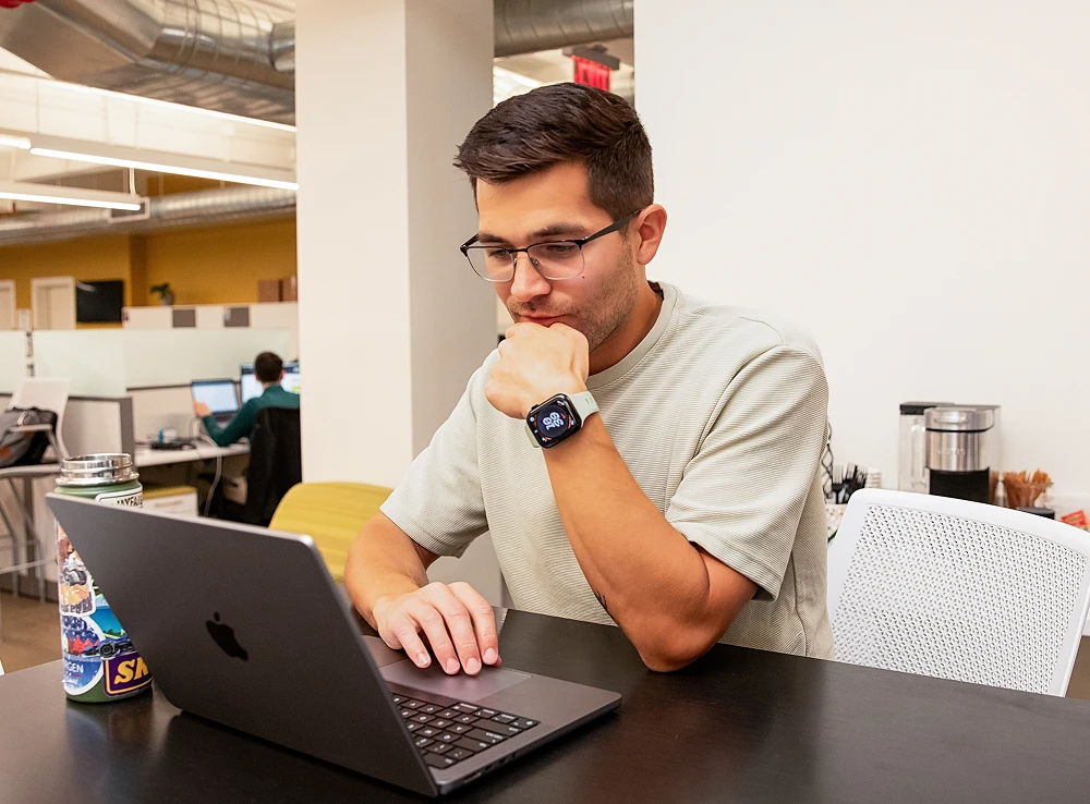 Young man wearing glasses focused on laptop at office desk with water bottle beside him.