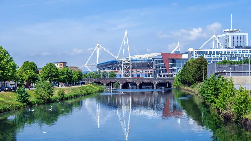 River with trees on both sides and a bridge with stadium and buildings in the background under blue sky.